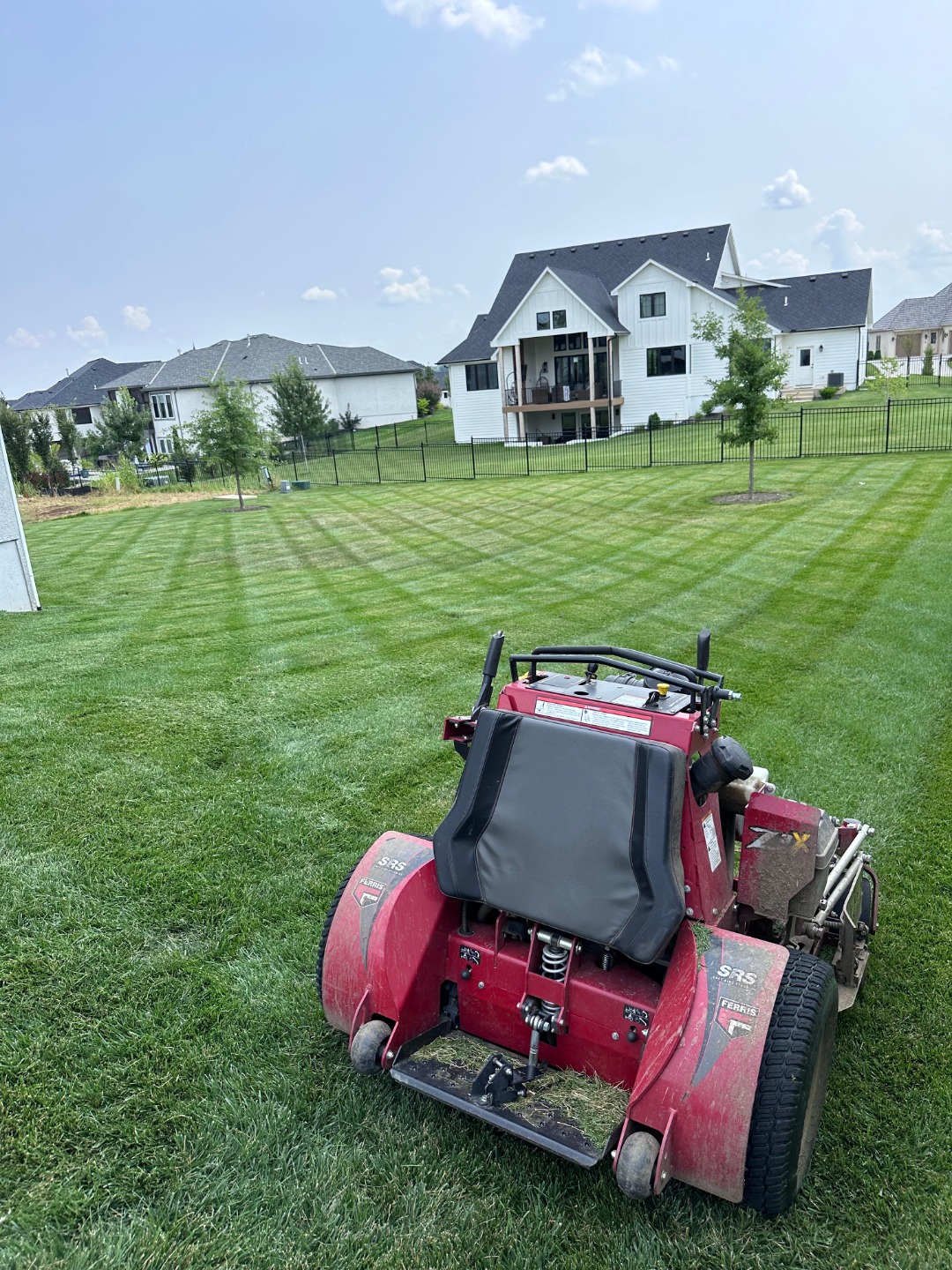 Lawn mower finishing a backyard with checkerboard mowing pattern near white modern homes and black fencing.